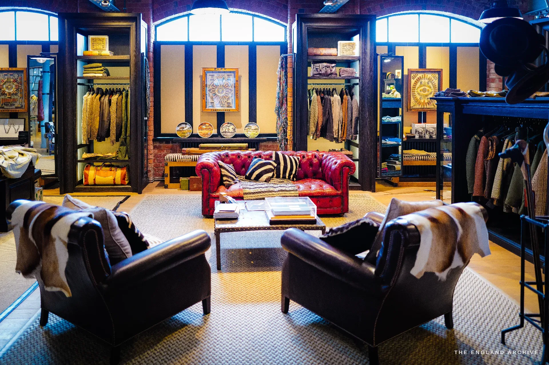 The sporting-luxury showroom: a red buttoned-leather Chesterfield sofa and two hide-draped armchairs around a low table, glazed display cabinets, tweed and safari clothing on rails, Eley cartridge display boards and arched windows in exposed brick.