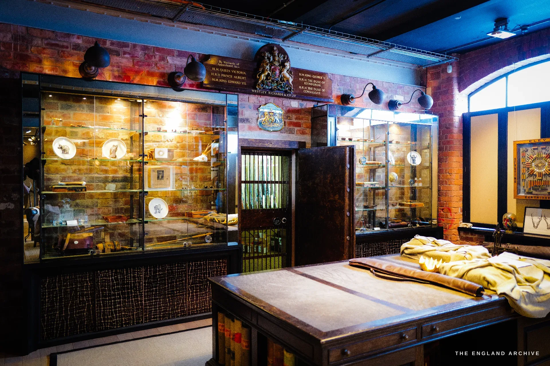 A heritage wall of glazed display cabinets flanking a steel strongroom door that stands open on a rack of guns; above, painted boards list royal warrants of appointment to Queen Victoria, Edward VII, George V and others, beneath the gilt royal arms.
