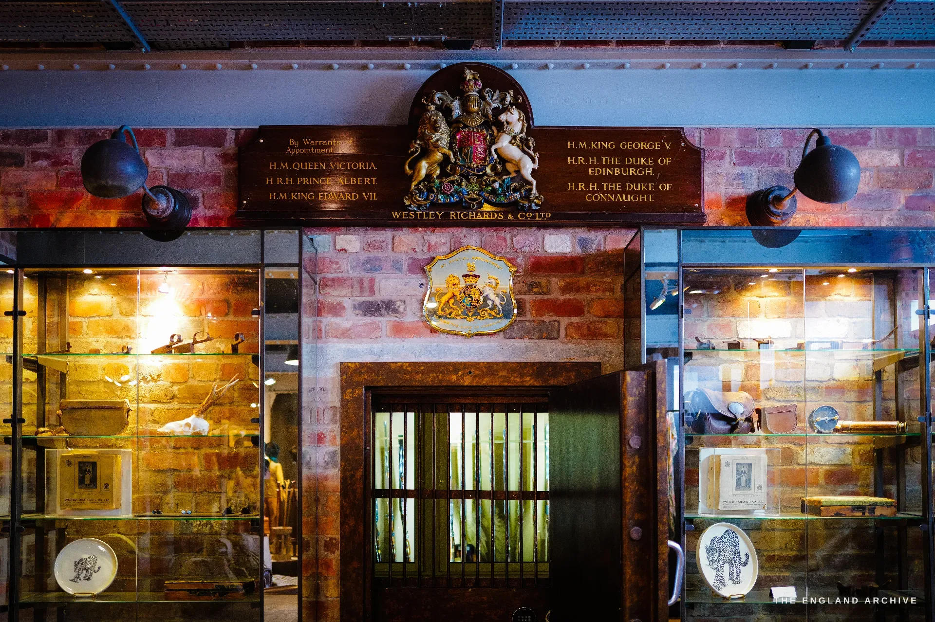A close view of the royal-warrant wall: painted boards naming appointments to Queen Victoria, Edward VII, George V and the royal dukes flank the gilt royal arms above a barred strongroom door, glazed display cabinets on either side in warm light on exposed brick.