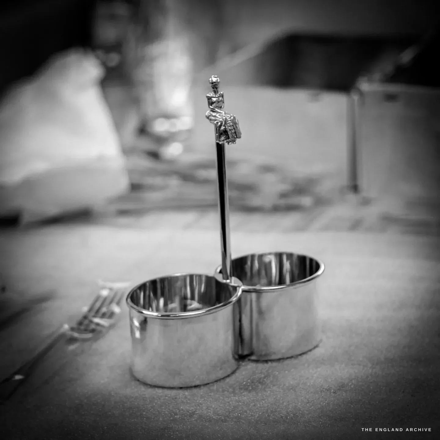 A finished silver twin-pot cruet stand with a slim central stem topped by a small cast figure, standing on a workbench.