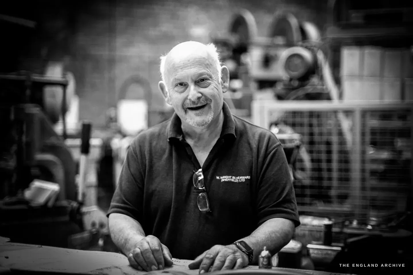 Stephen Wright leaning on a workbench, smiling broadly to camera, sunglasses hooked on the collar of his firm polo shirt, machinery behind him.