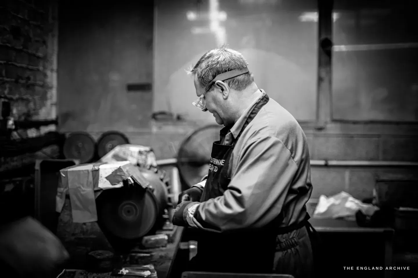 A worker in safety glasses and a William Wright apron holding a piece to a spinning buffing mop, the wheel blurred with motion.