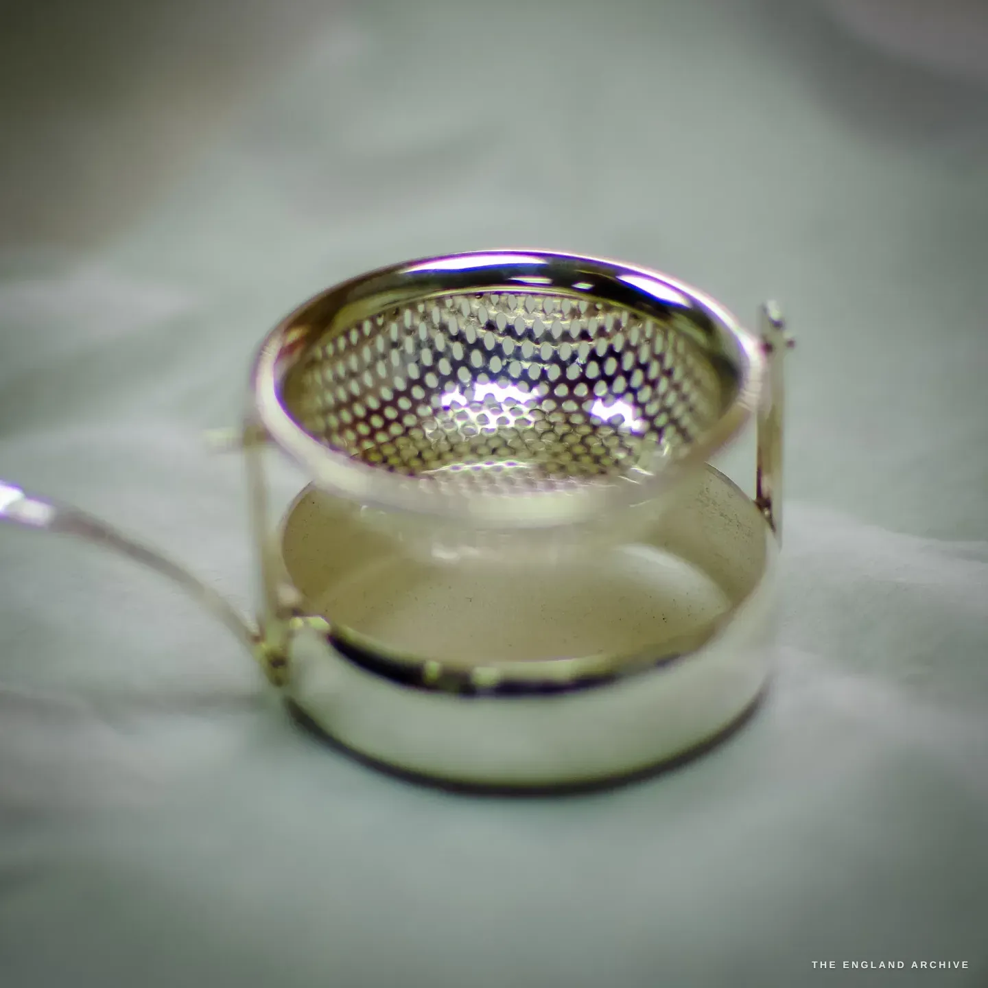 A finished silver tea strainer, its pierced mesh bowl held in a ring above a small drip bowl.