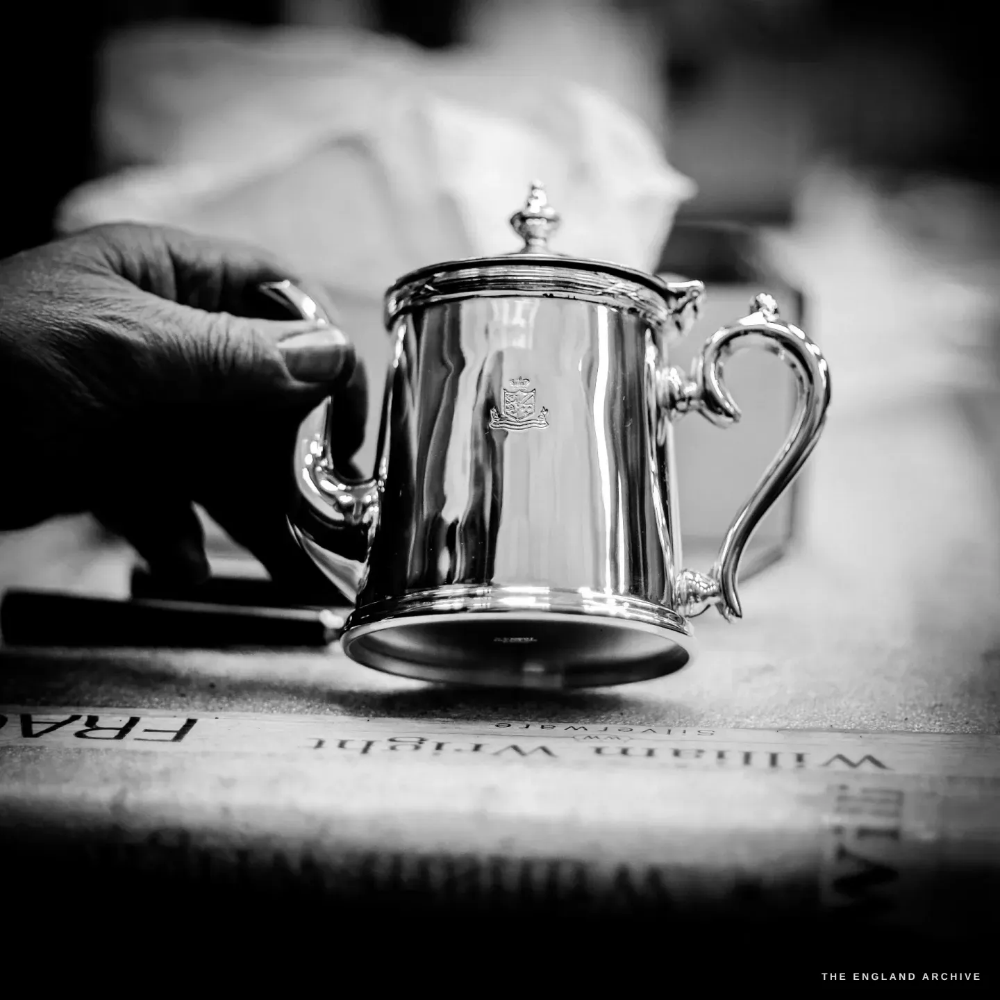 A small crested silver lidded pot with an ornate handle, steadied by a hand, standing on a packing box printed William Wright Silverware.