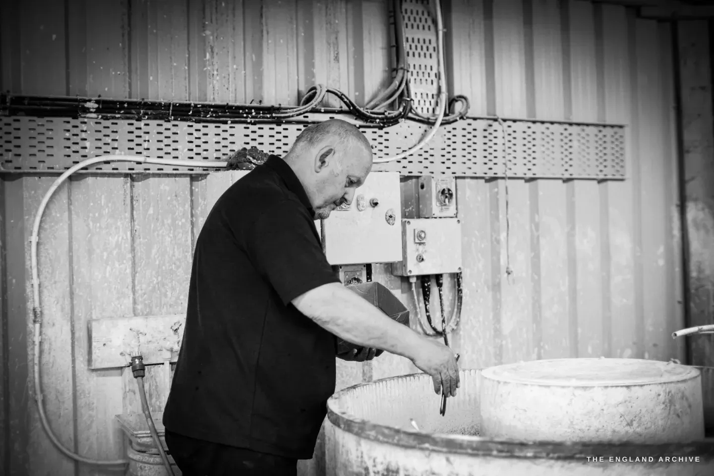 Stephen Wright leaning over a large round barrel against a corrugated wall, lowering work down into it.