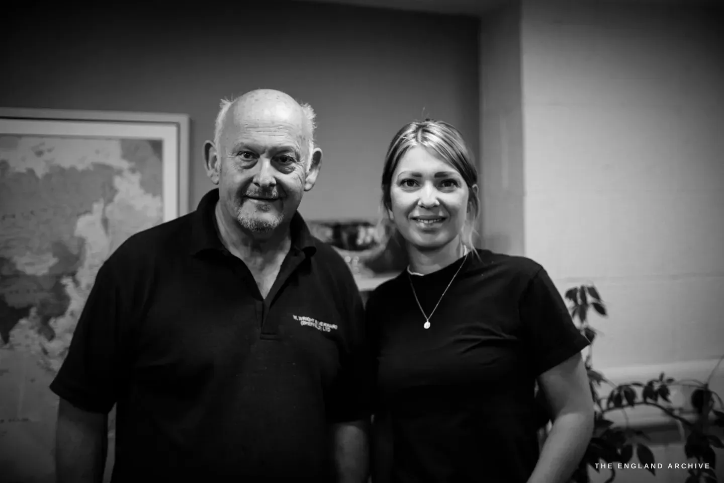 Stephen Wright and his daughter standing together in the office, smiling to camera, a framed world map on the wall behind them.