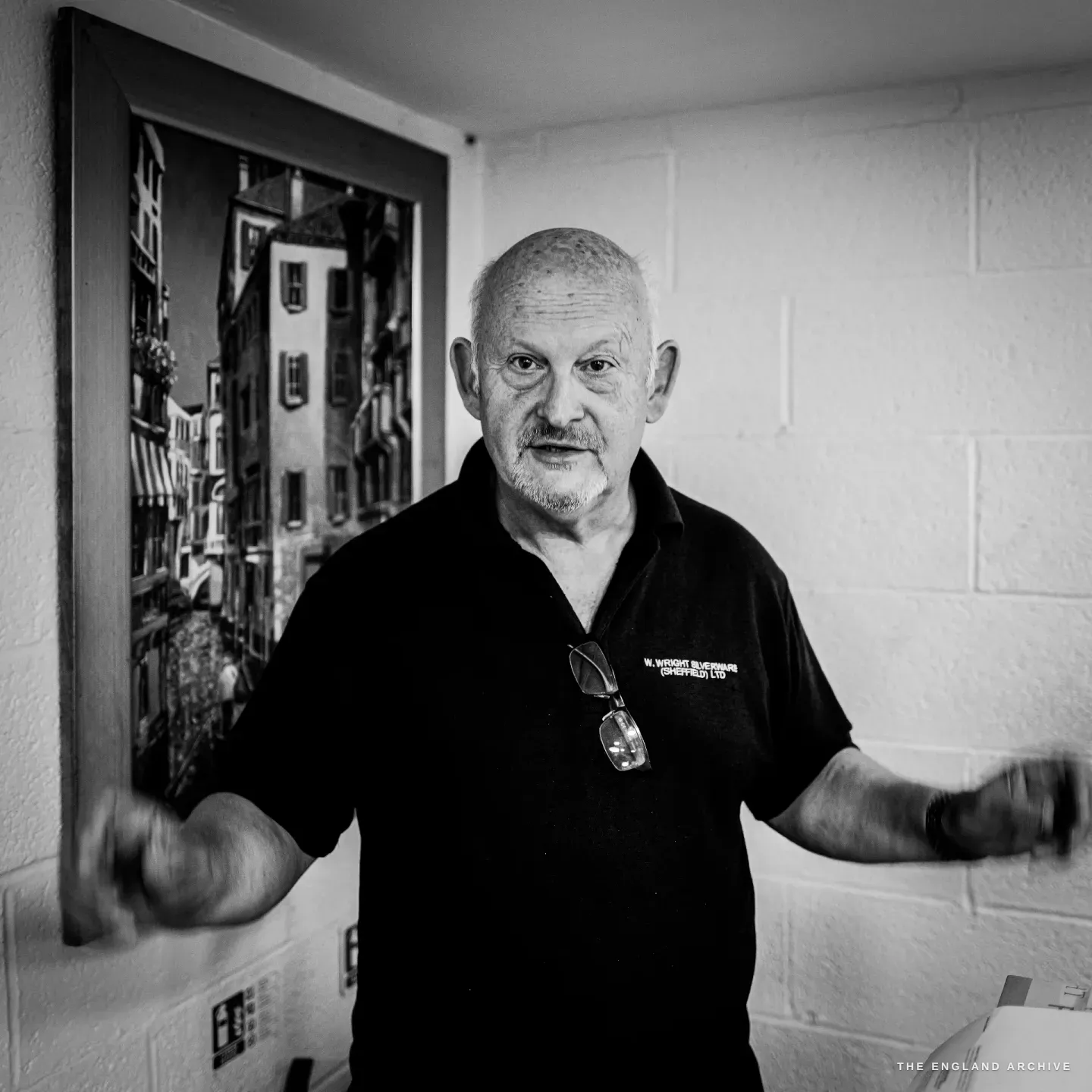 Stephen Wright talking with his hands blurred in motion, beside a framed photograph of a European old-town street on a whitewashed wall.