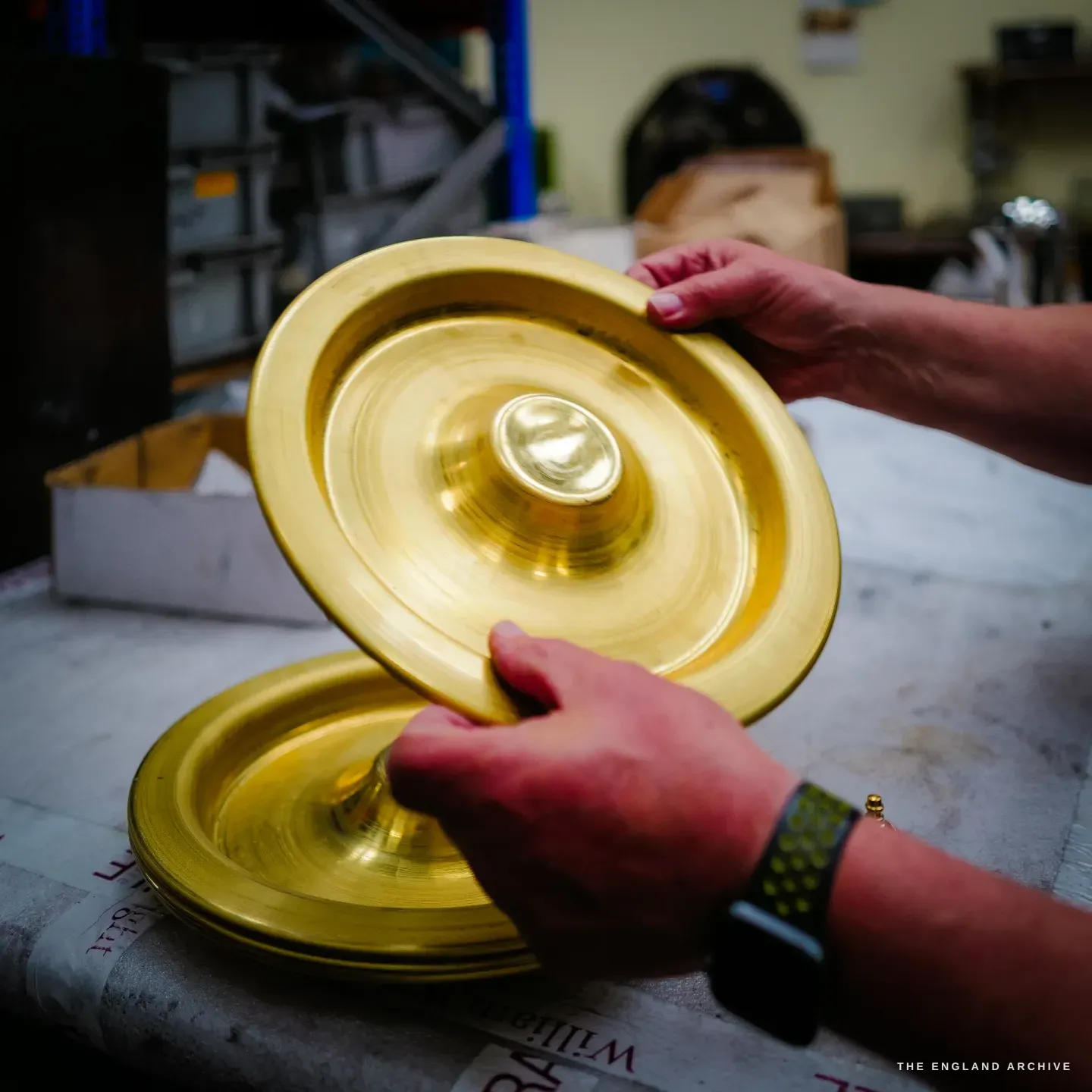 Hands holding two spun brass dishes, the raw gold-coloured metal bright before it is plated, on the William Wright bench paper.