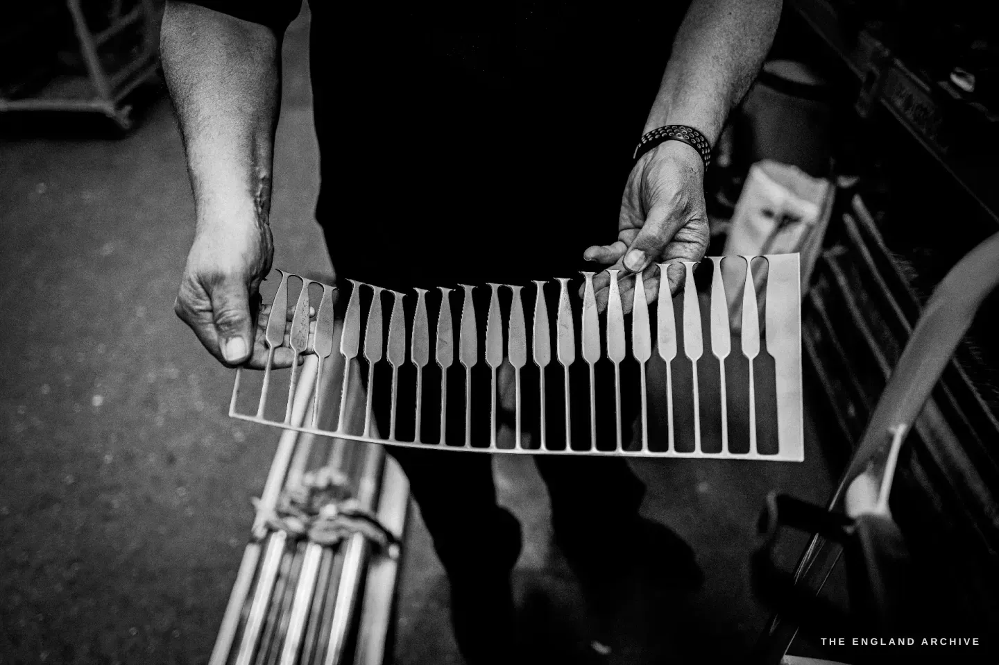 Stephen Wright’s hands holding a flat strip of stamped spoon blanks, still joined in a row to the parent sheet of metal.