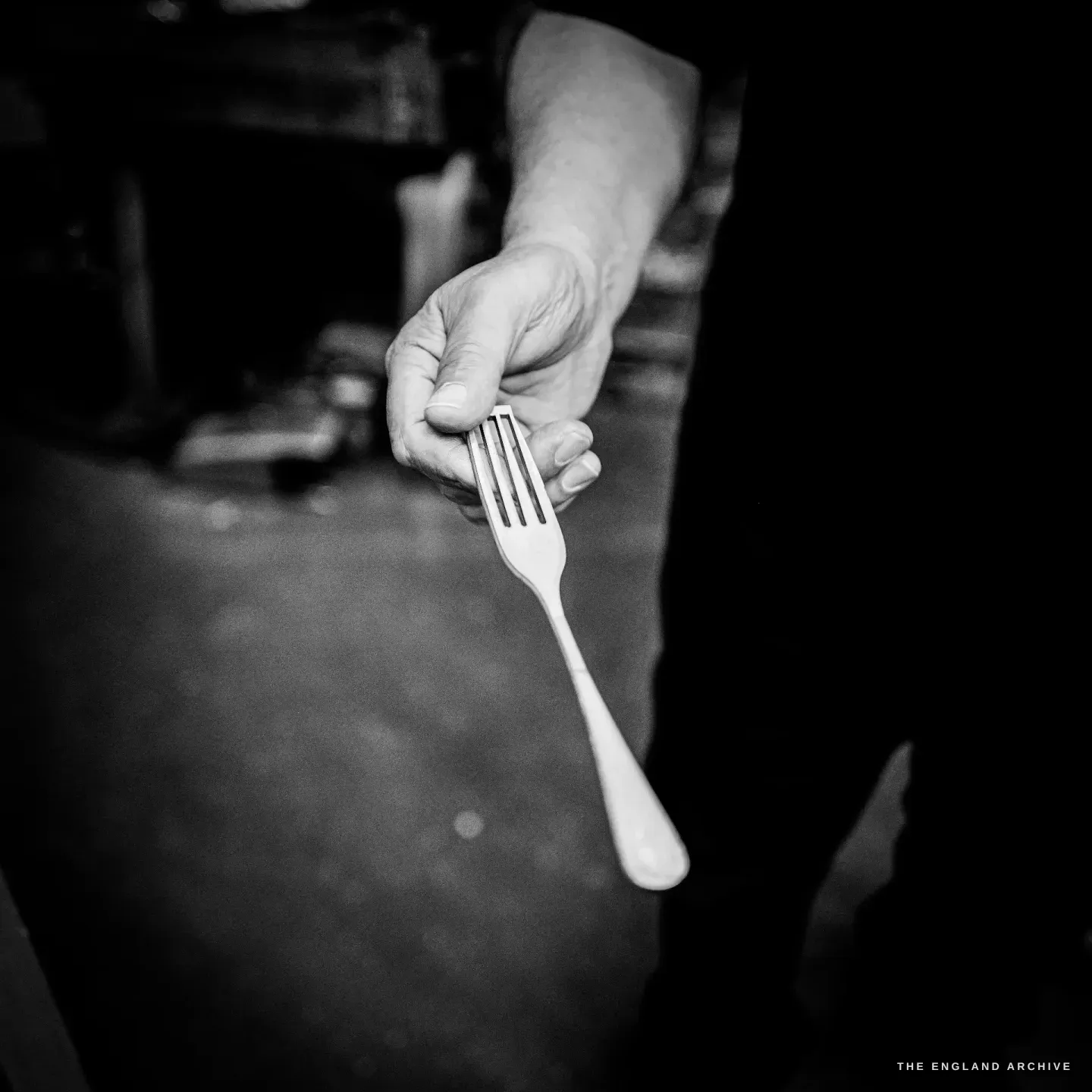 Stephen Wright’s hand holding out a single finished table fork toward the camera against a dark background.