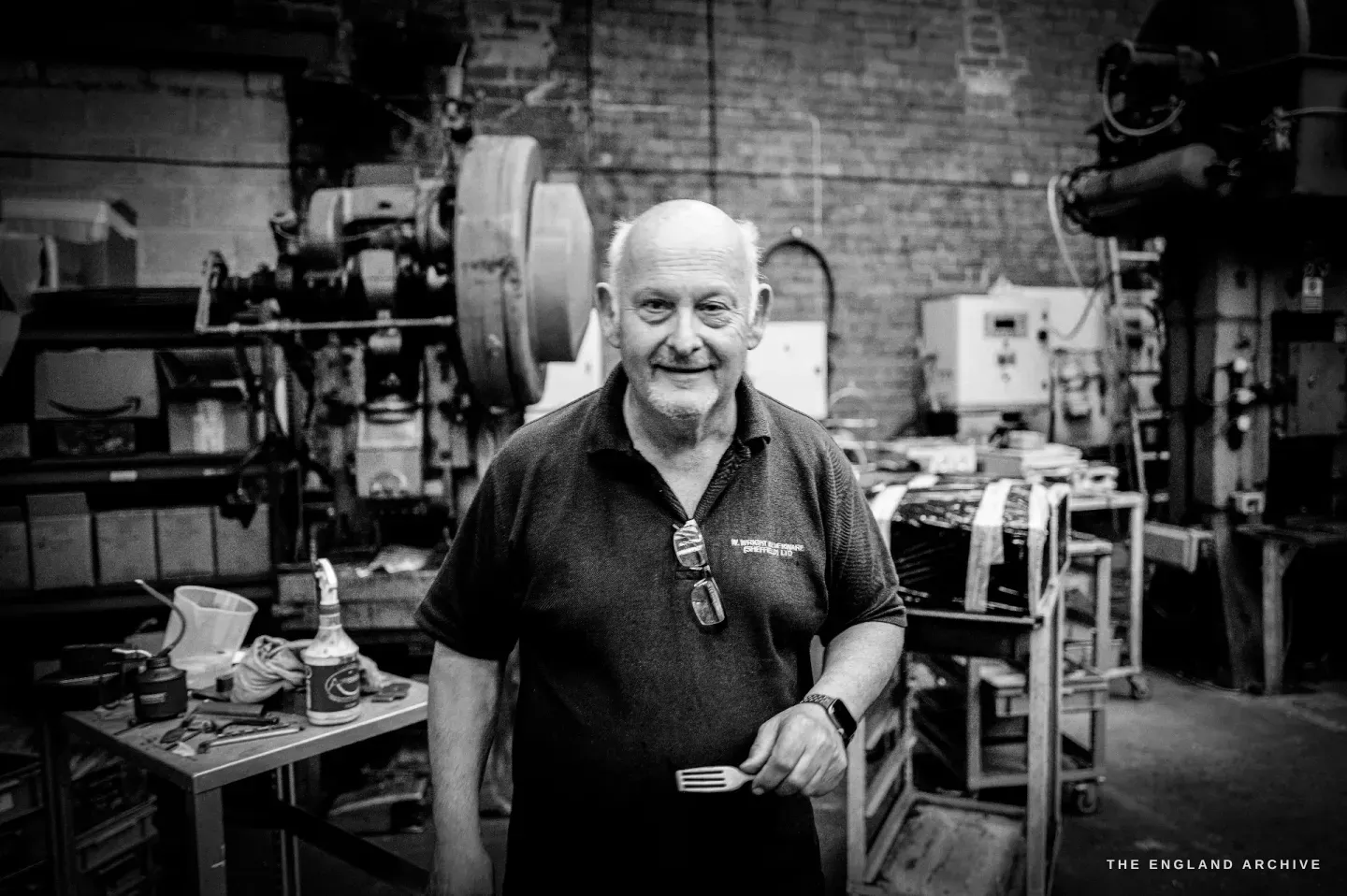 Stephen Wright standing among the drop-stamp presses of his workshop, smiling, a finished fork in his hand.
