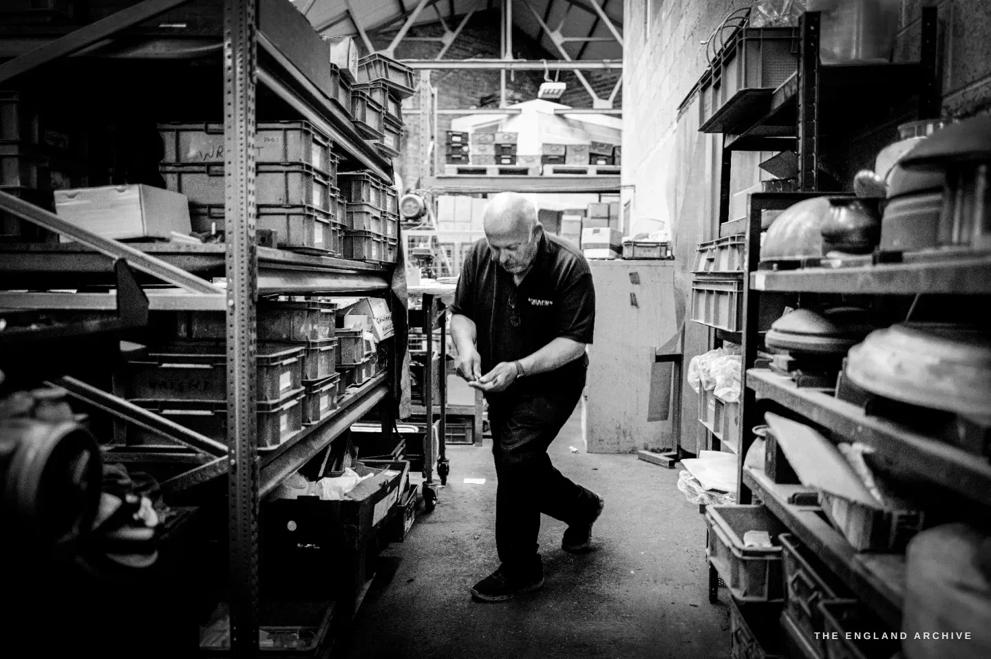 Stephen Wright walking a narrow aisle between tall racks crammed with parts bins, tools and stacked steel forms.