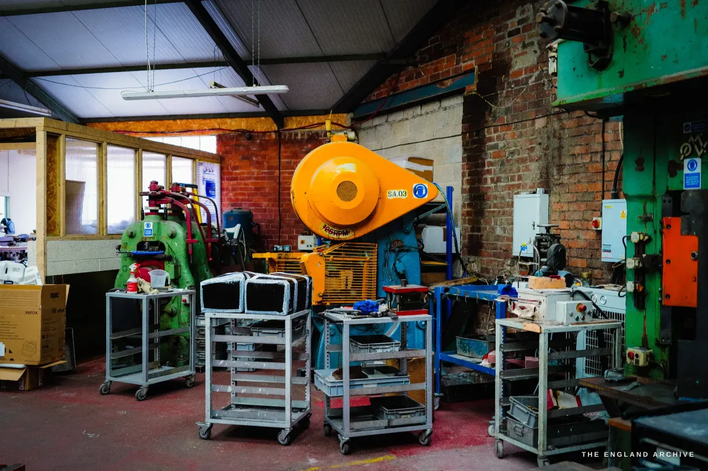 A wide view of the press shop in colour, an orange power press, a green machine and red-painted machinery, trolleys of dies between them, a brick wall and red floor.