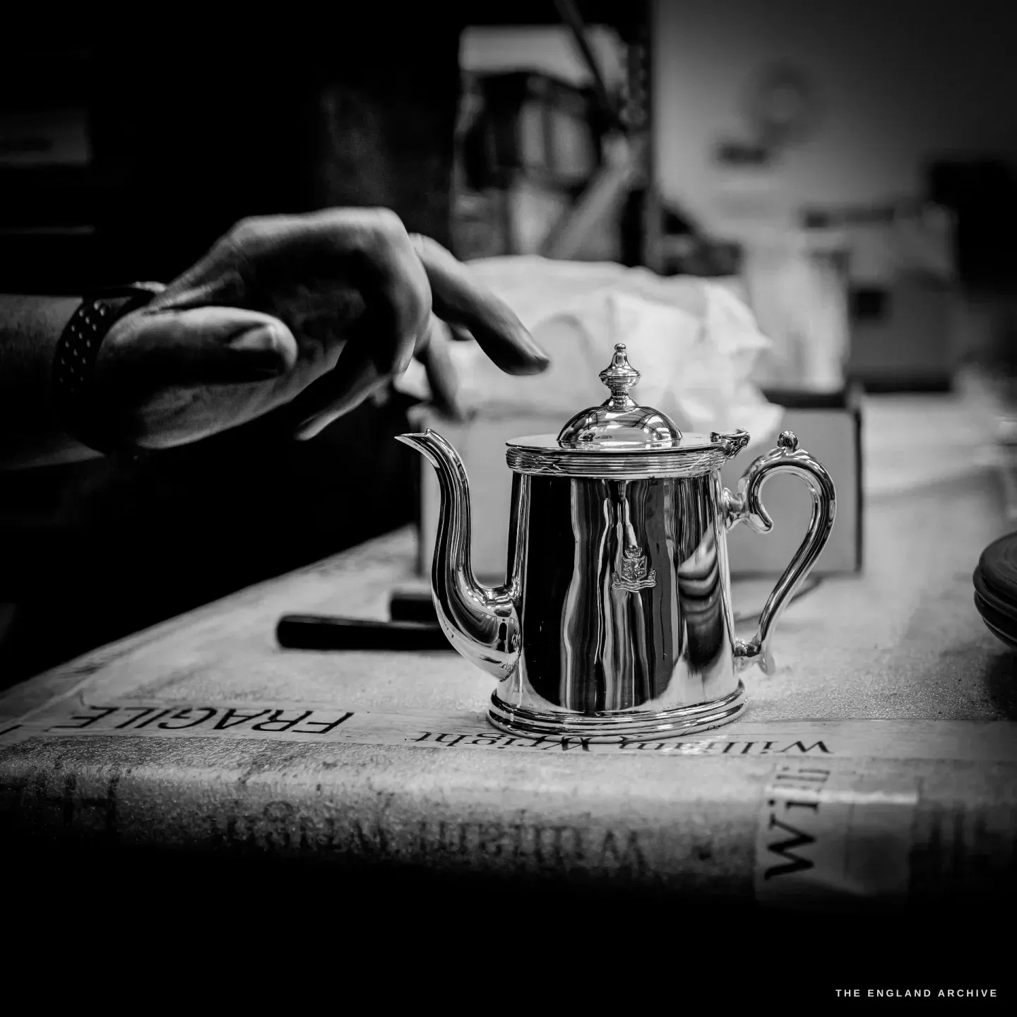A finished crested silver teapot with an ornate handle and finial lid standing on a packing box printed William Wright and Fragile, Stephen Wright’s hand gesturing toward it.