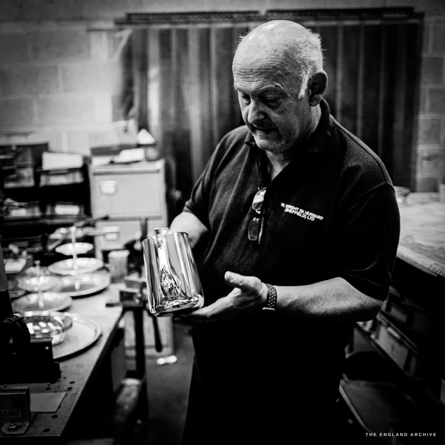 Stephen Wright holding and inspecting a finished silver milk jug in both hands, a row of finished dishes on the bench beside him.