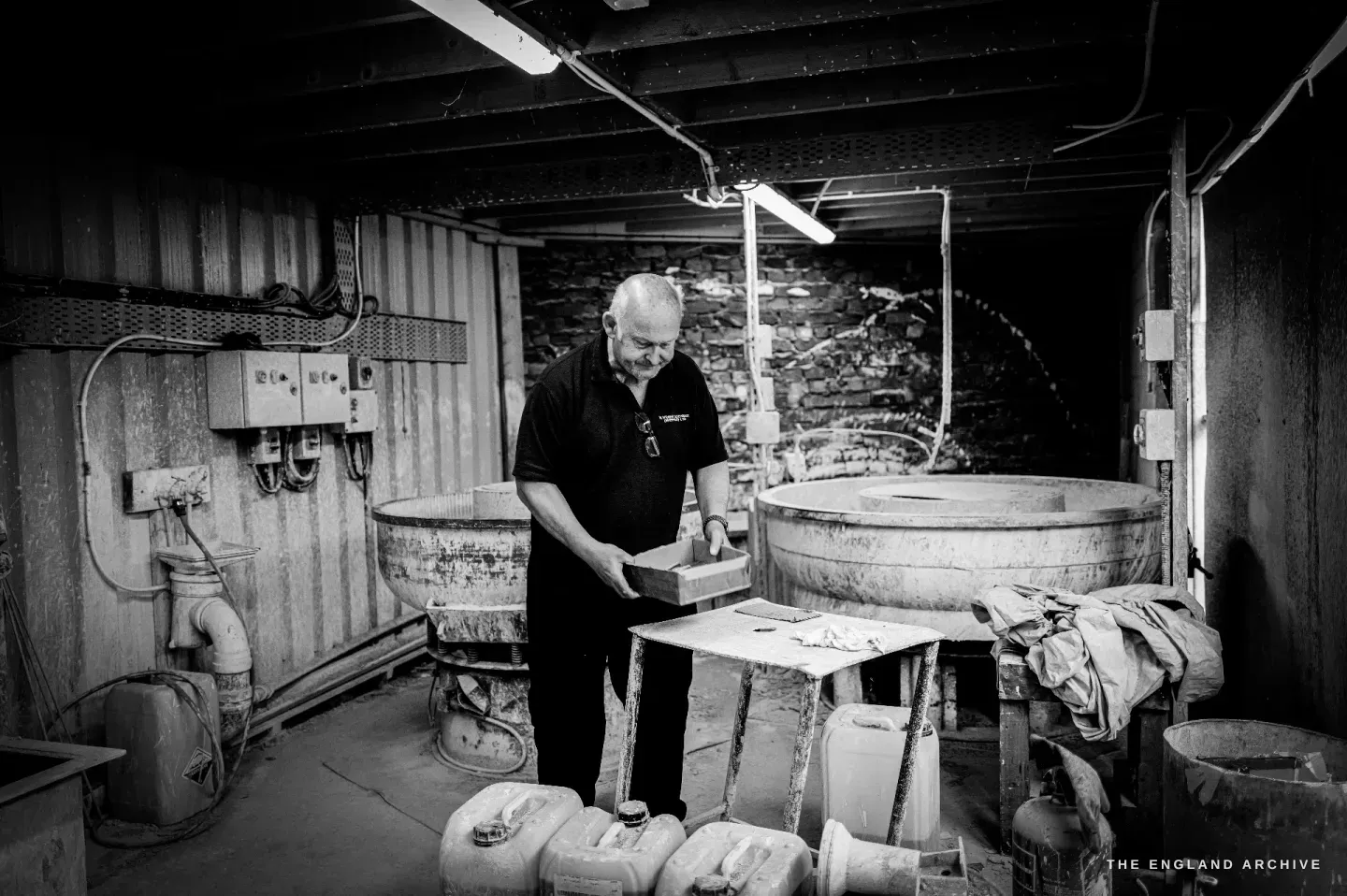 Stephen Wright in the plating room among large round tubs and drums of chemicals, holding a tray, a stone wall behind.