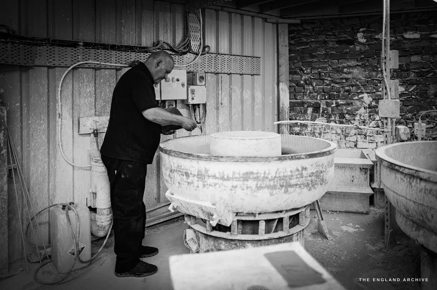 Stephen Wright at a round vibratory finishing machine, a stone wall behind him, working over the open bowl.