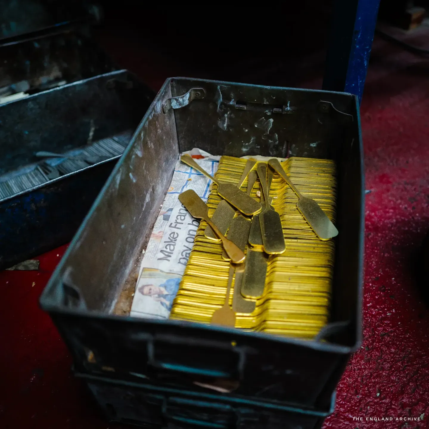 A steel crate filled with stacked golden brass spoon blanks on newspaper, a red floor beyond.