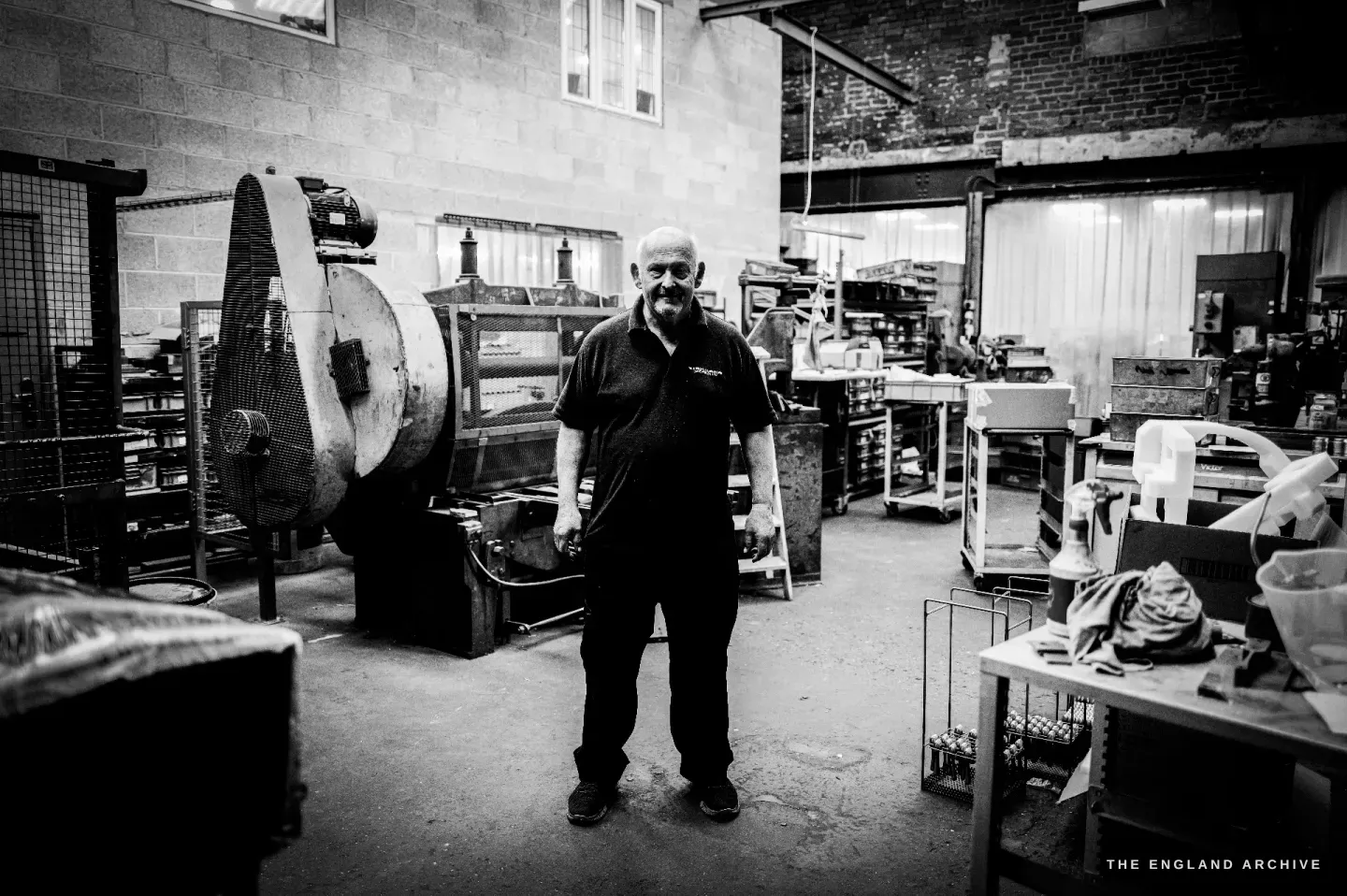 Stephen Wright standing full-length in the centre of his workshop, the round finishing barrel to his left and benches around him.