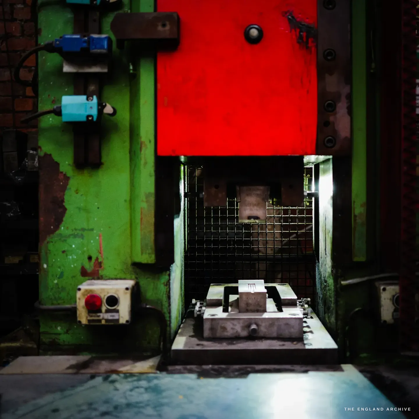 A power press seen head-on, a bright red ram over a green frame, a die set on the bed behind a mesh guard, brick wall and blue electrical boxes.