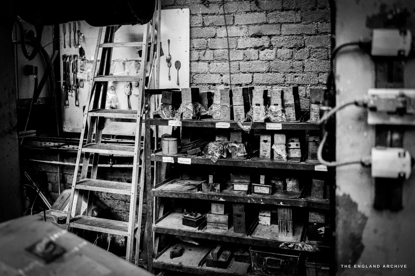 A storage corner with a wooden stepladder, shelves stacked with steel dies, and cutlery pattern templates hung on the brick wall.