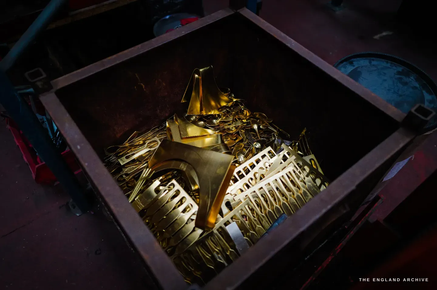 A steel bin filled with golden brass blanking scrap, the lattice skeleton left after blanks are stamped from sheet, glowing in the dark.