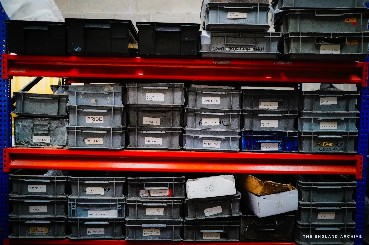 Red-railed industrial racking of grey parts trays, each hand-labelled with a cutlery pattern name including PRIDE and QUEENS.