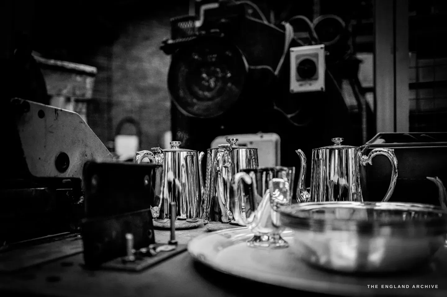 A row of finished mirror-polished silver teapots and jugs gleaming on a bench, a footed dish and a bowl in front, dark press machinery behind.