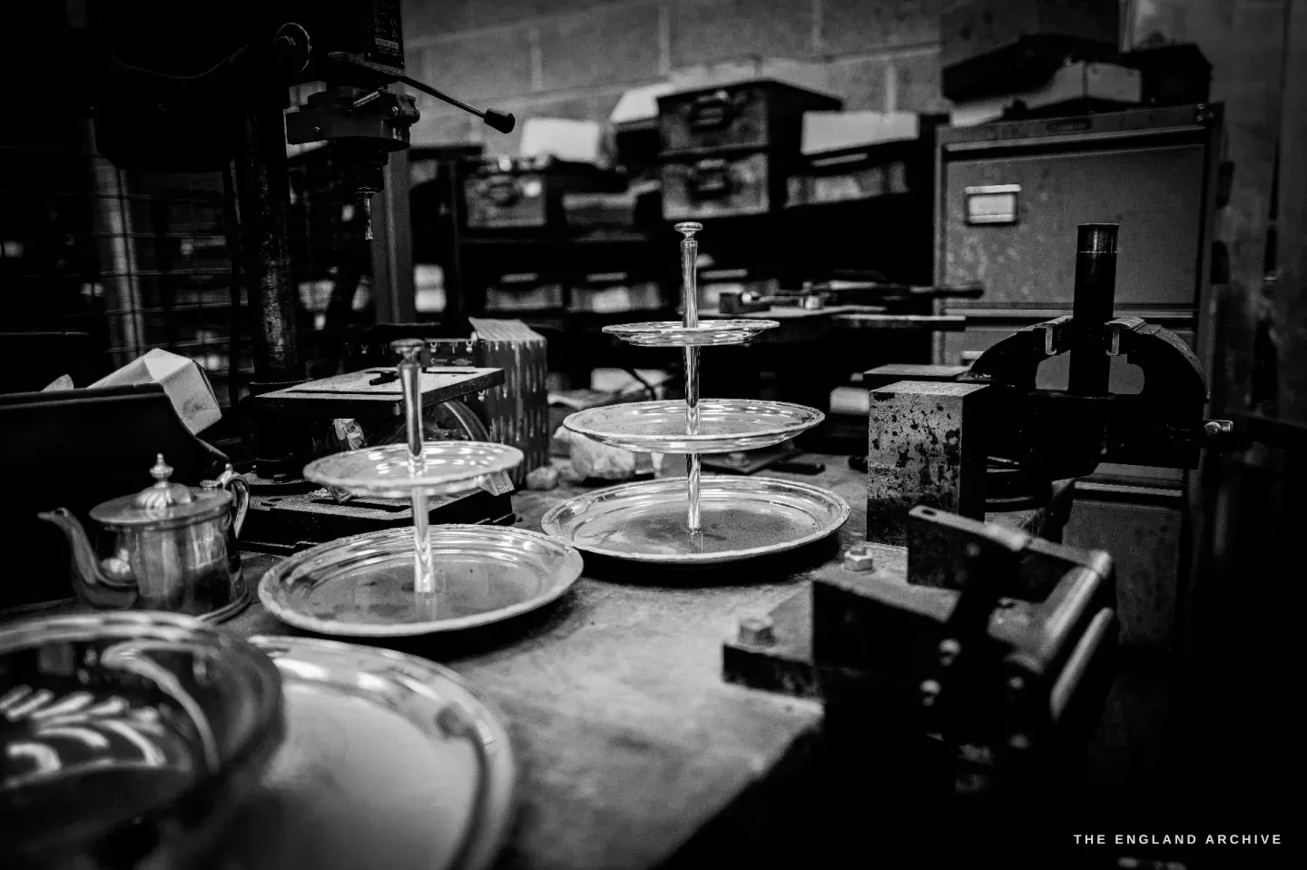 Finished silver tiered cake stands, round trays and a teapot grouped on a workshop bench.