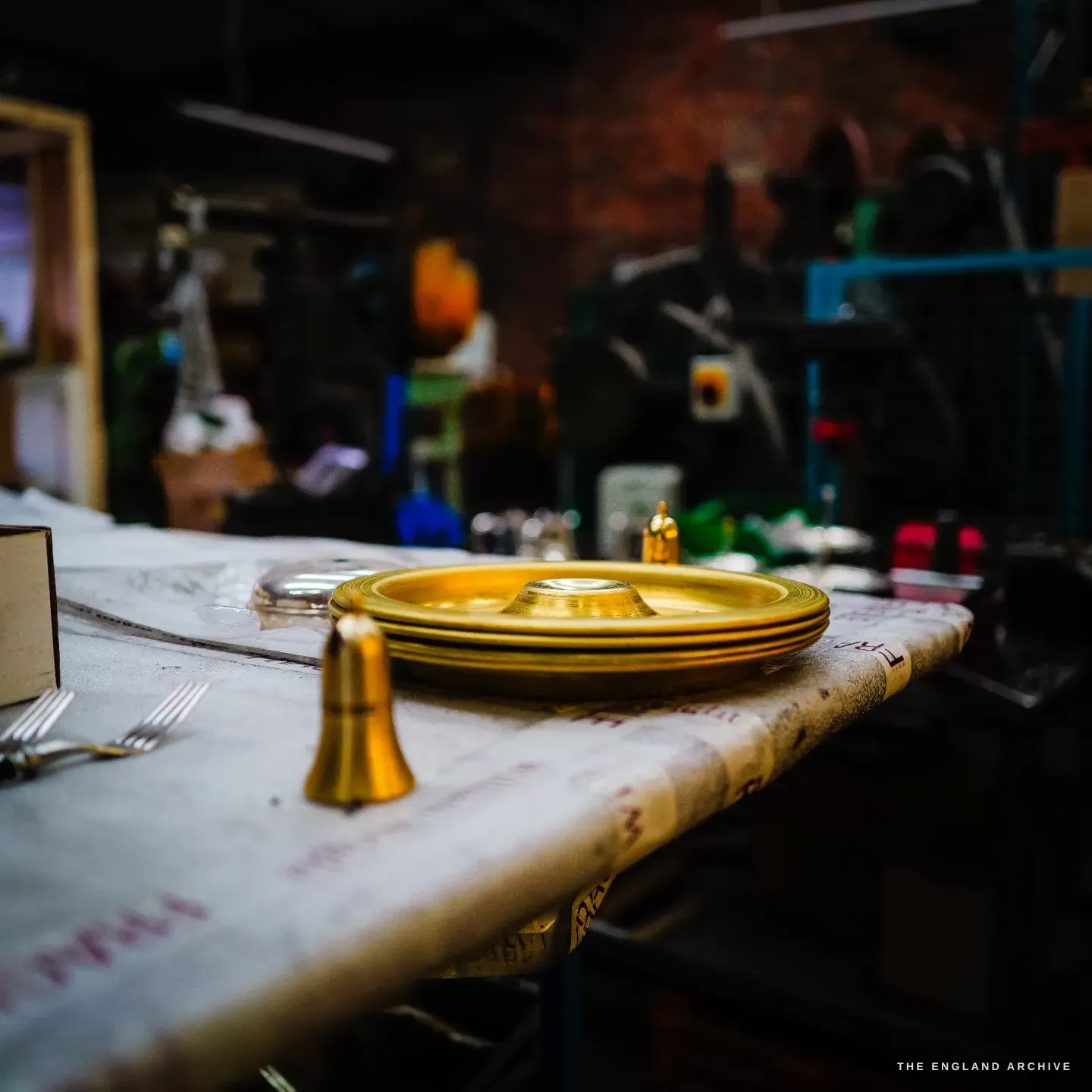 Golden brass spun dishes stacked beside a brass caster body on the paper-covered bench, silver forks at left, dark brick behind.