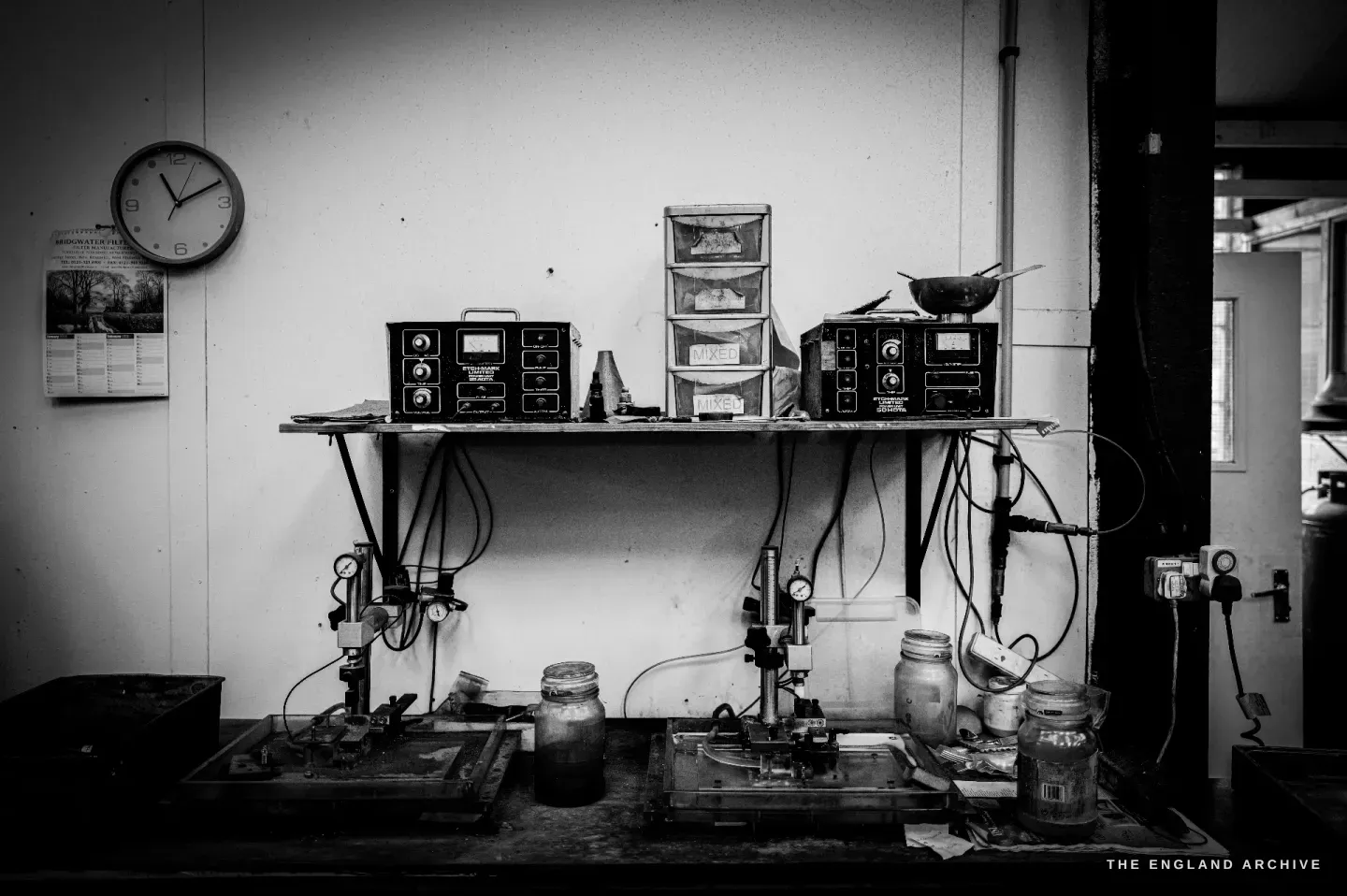A soldering and plating bench with two torch setups, jars and control boxes, a clock on the wall.