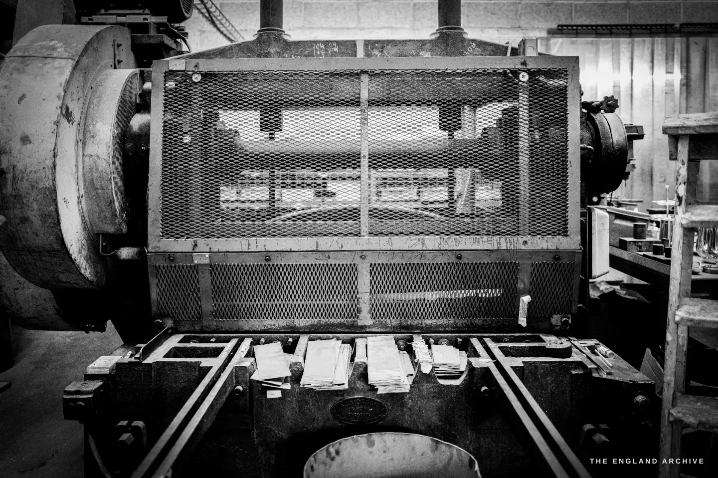A large press seen head-on behind a mesh guard, a heavy flywheel at left and dies and stacked blanks in the bed.