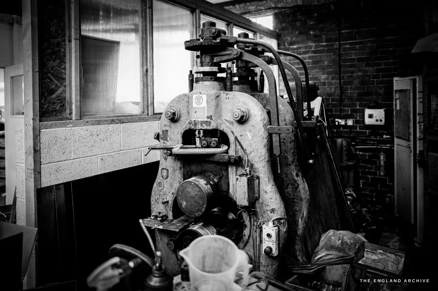 A large worn drop-stamp press seen head-on, a maker’s plate on its frame, an office window to the left.