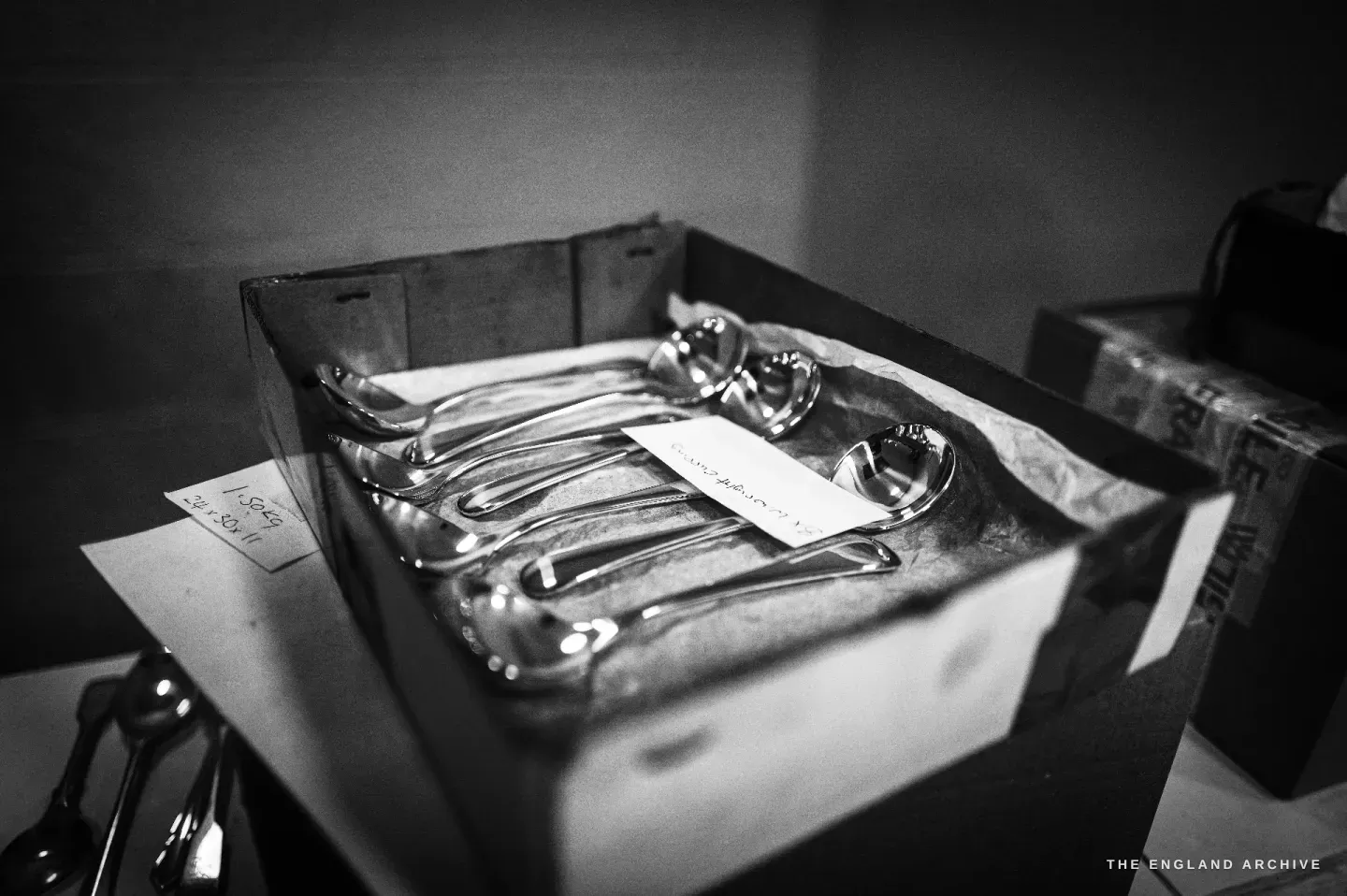 A box of finished silver ladles and serving spoons laid in tissue with a handwritten note, a Fragile box to the right.