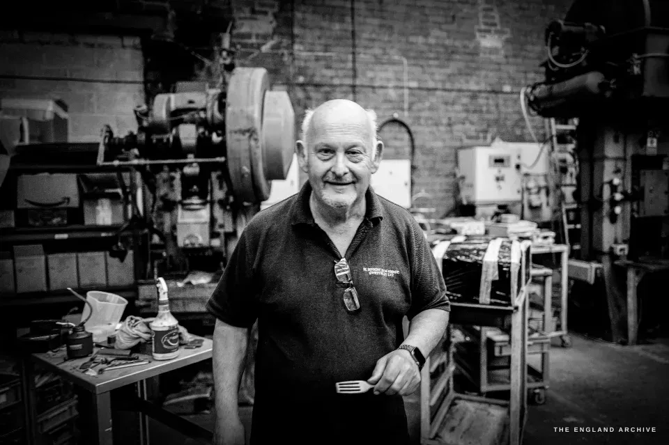 Stephen Wright standing among the drop-stamp presses of his workshop, smiling, a finished fork in his hand.
