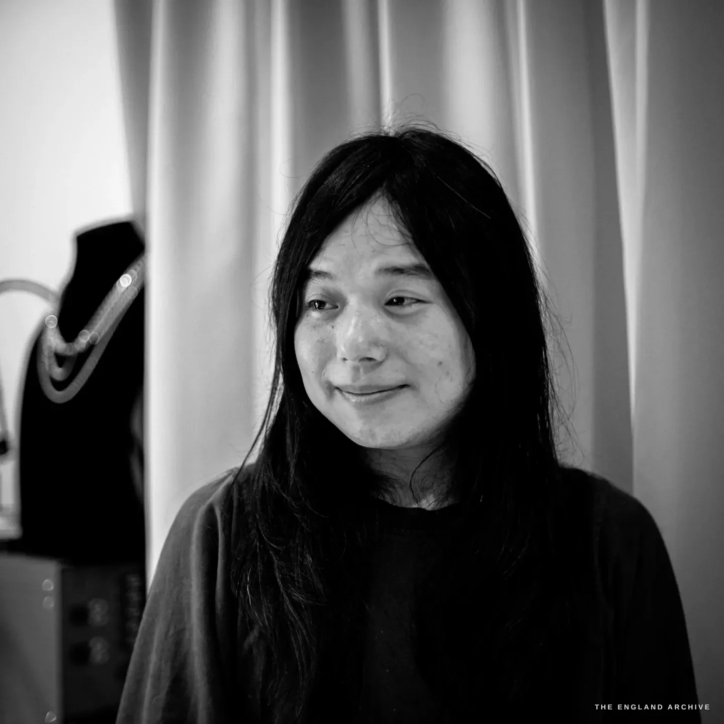 A calm, direct portrait of Yuyi Cheng, dark hair down, a soft smile, a draped necklace bust just visible at the edge of the frame.