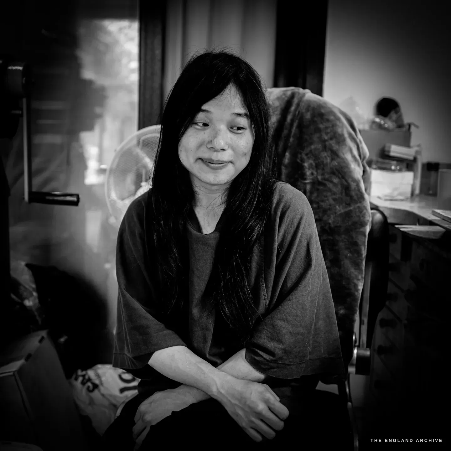 Yuyi seated in a chair beside her bench, arms resting on her lap, an electric fan and bench drawers behind.
