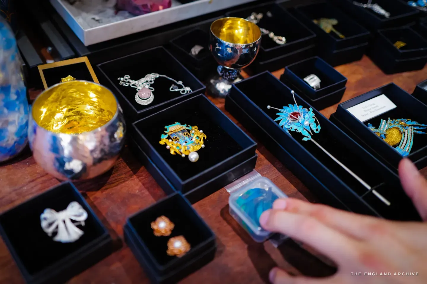 Overhead view of finished pieces in black display trays on a wooden table: a gold-lined silver cup, a rose-quartz filigree pendant, a blue-and-gold feather brooch with pearls, a feather hairpin, pearl flower studs and a mother-of-pearl bow, with a hand reaching in holding a small box of loose blue feathers.