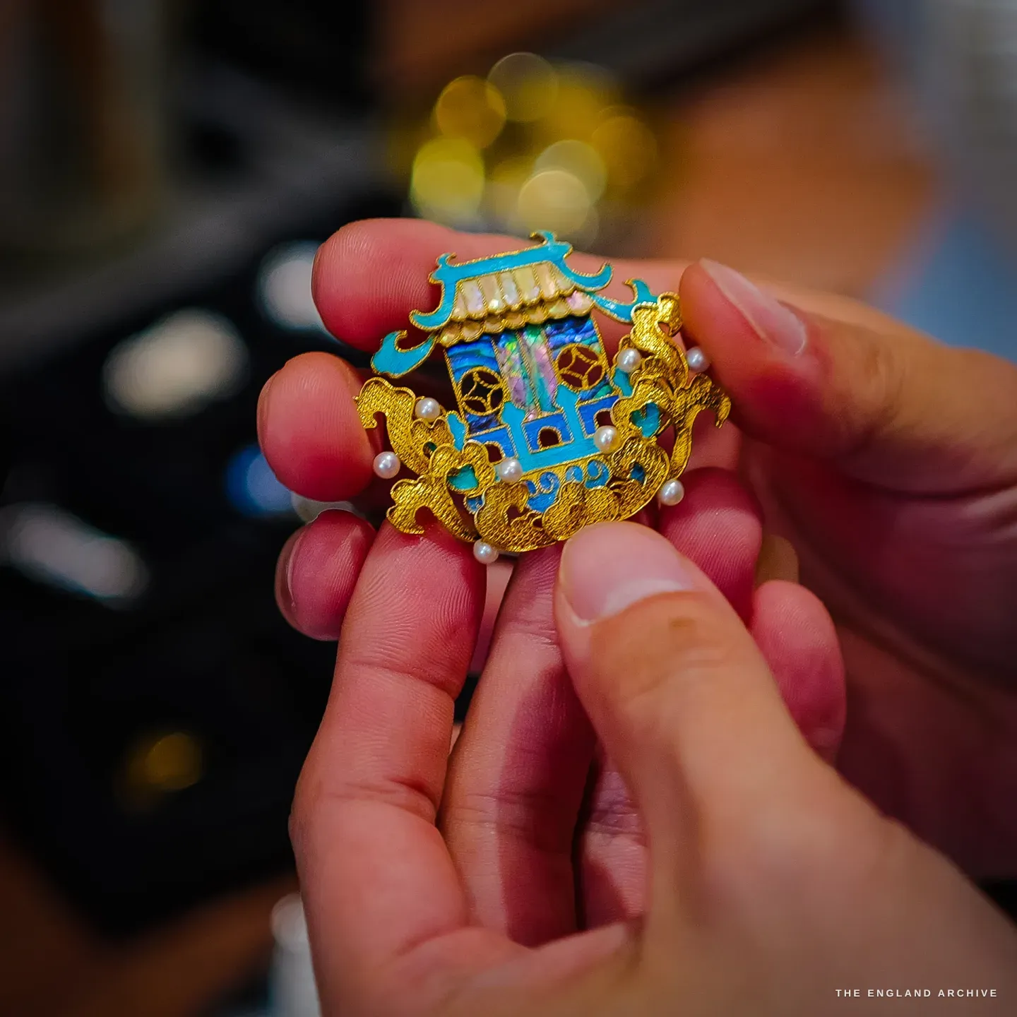 A hand holds a gold-filigree brooch shaped like a pavilion, its blue panels made of dian cui feather inlay, set with seed pearls and iridescent mother-of-pearl.