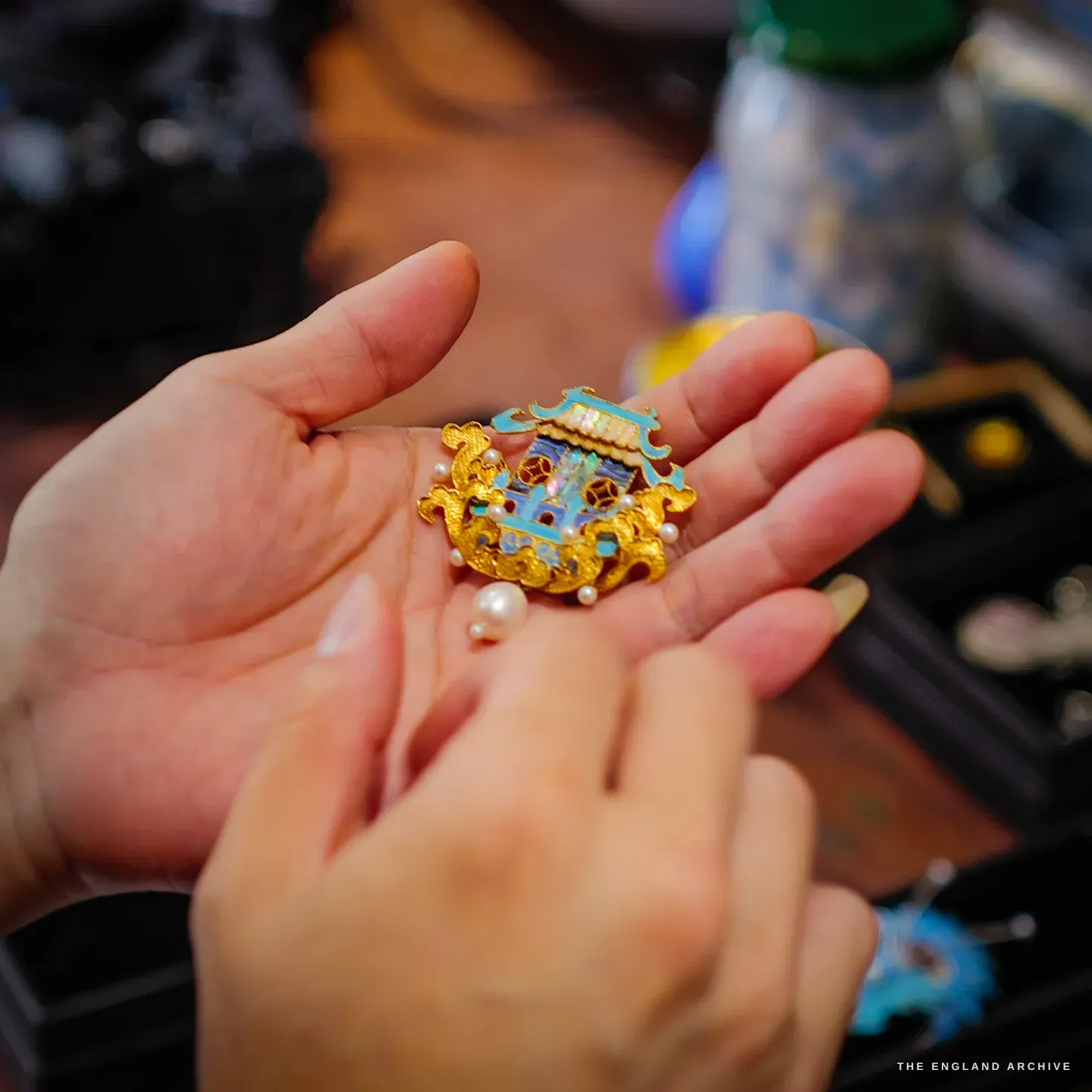 The same gold and blue pavilion brooch resting on an open palm, a loose pearl beside it, the studio soft behind.
