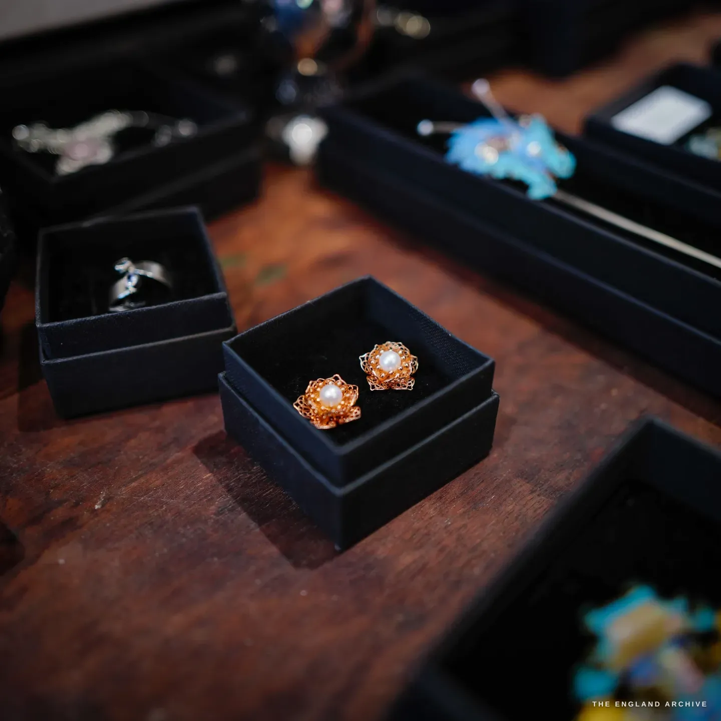 A black box of rose-gold filigree pearl flower stud earrings in the foreground, a silver ring and a blue feather hairpin in trays behind.