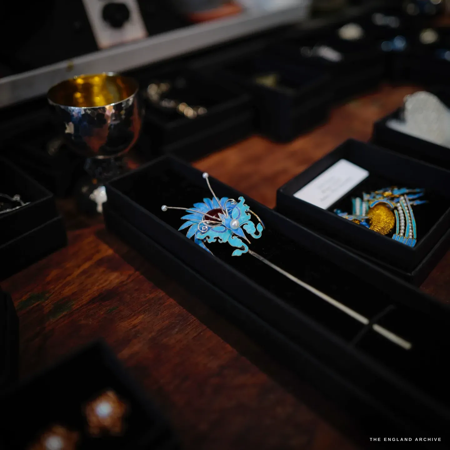 A blue feather and pearl hairpin with a red cabochon laid in a black tray beside a gold-lined silver cup.