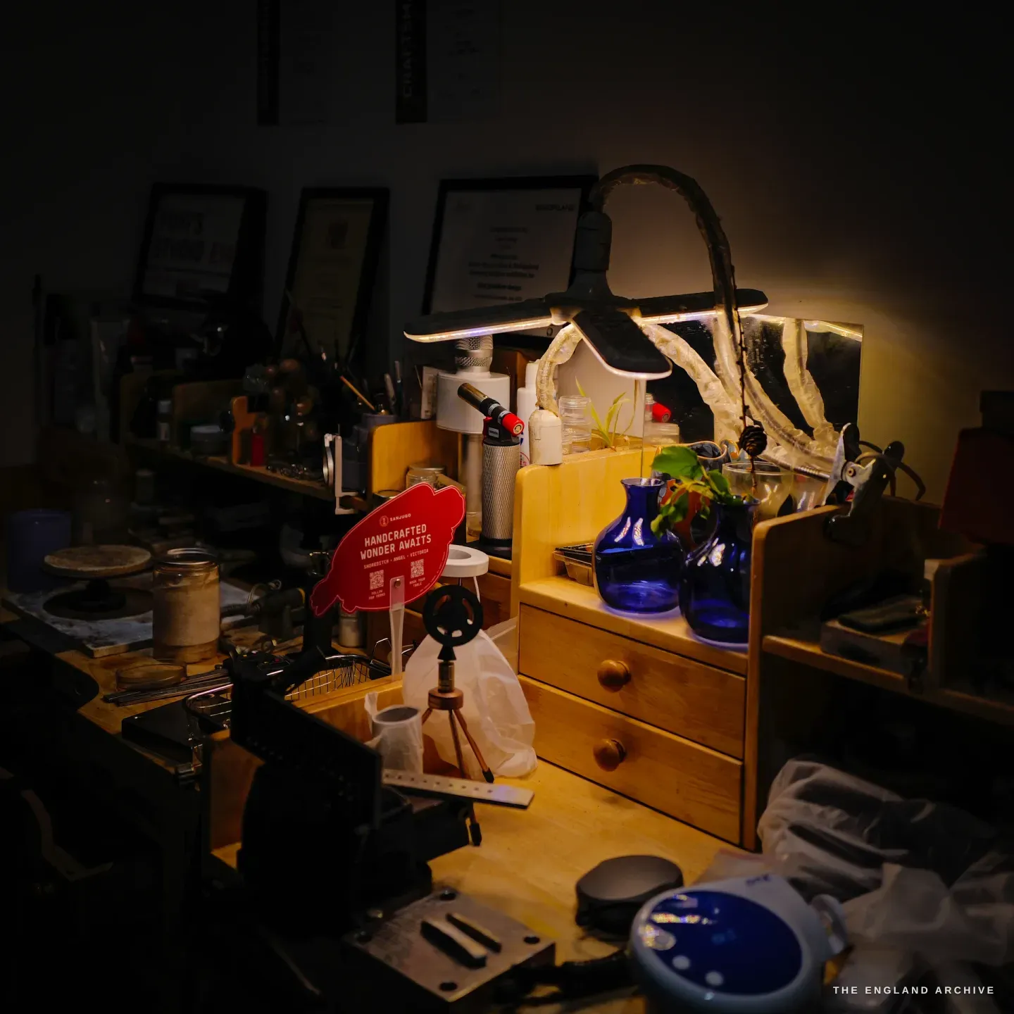 The workbench corner under an anglepoise lamp: a hand torch, jars, a wooden organiser with blue glass vases and a plant cutting, framed certificates on the wall above.