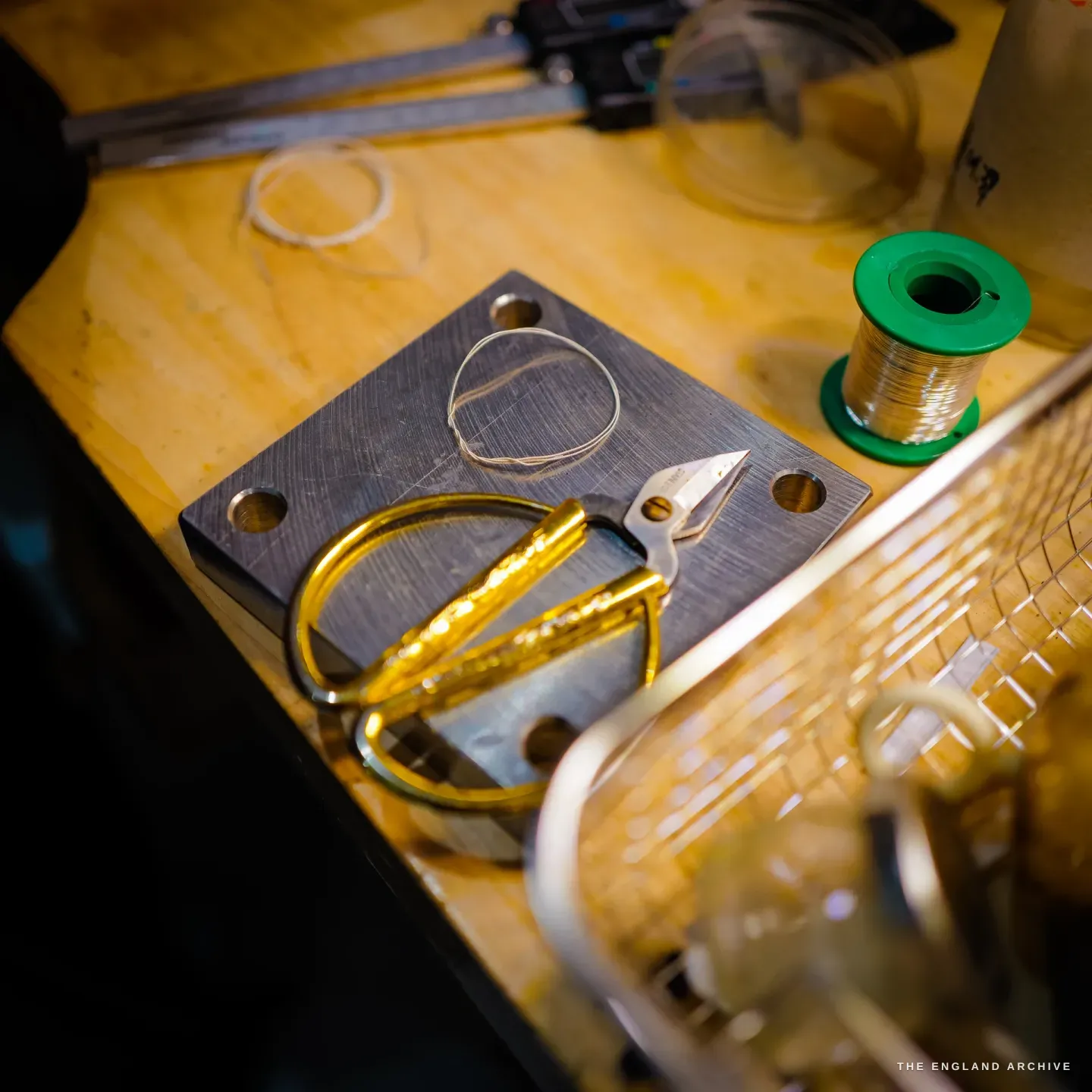 A bench detail: a steel drawplate block, gold-handled snips, a coil of silver wire, a green spool of fine wire and a pair of calipers.