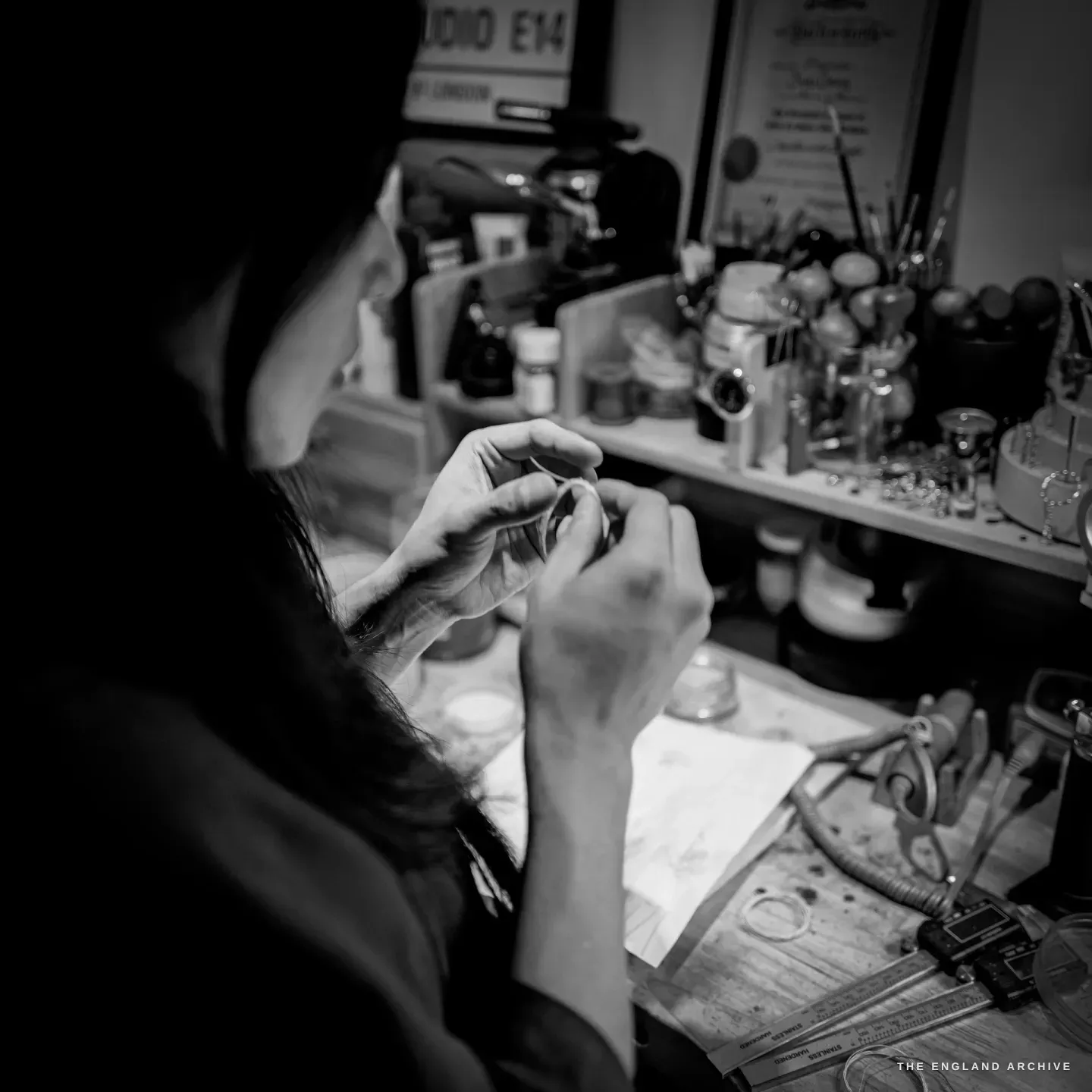 Over-the-shoulder of Yuyi’s hands manipulating a thin silver ring of wire at the bench, a shelf of pots and tools behind.