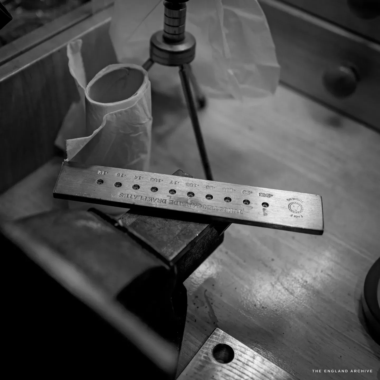 A tungsten-carbide drawplate clamped in a vice, stamped with wire-gauge figures, beside a torn paper roll.