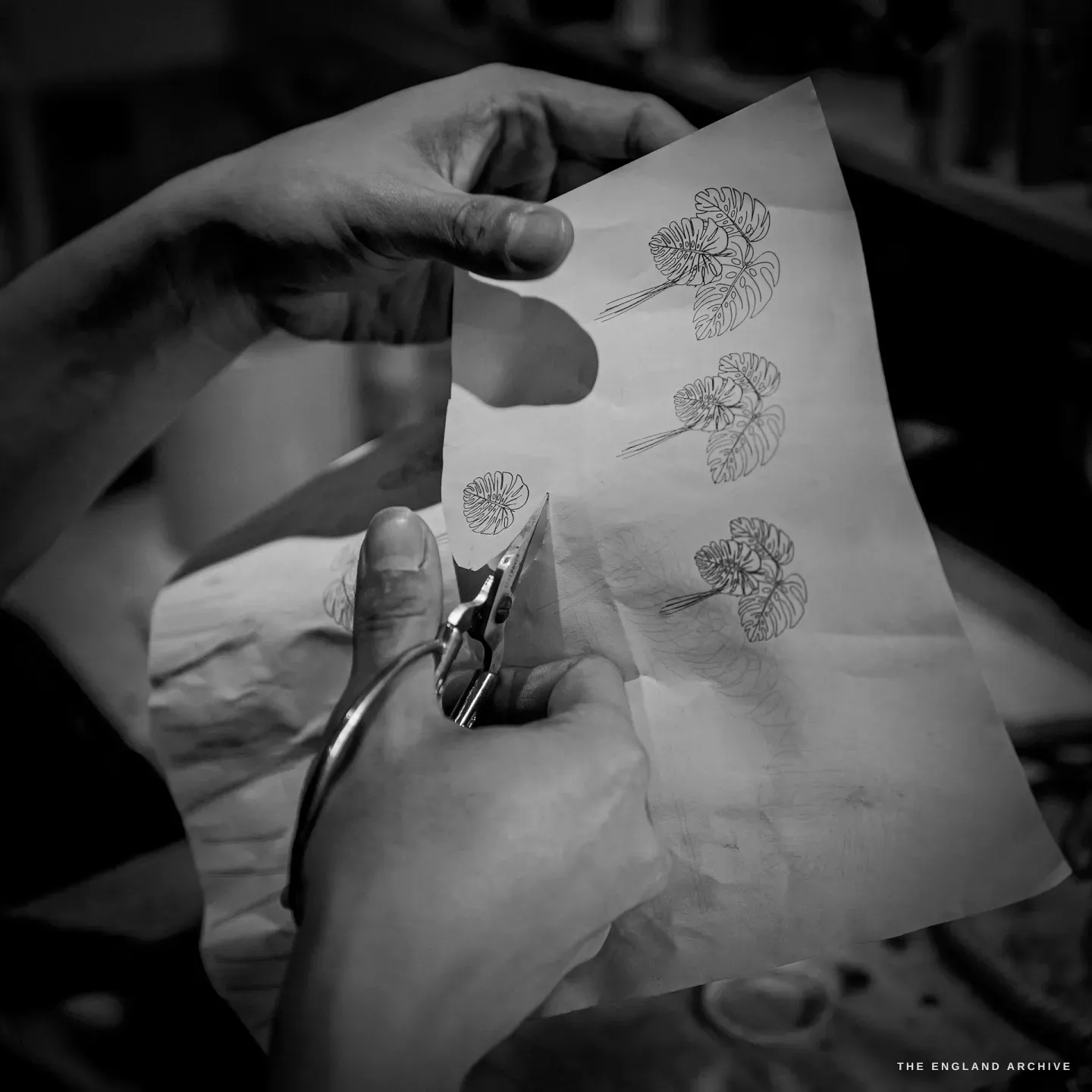 Two hands cutting out paper templates of monstera leaves with snips, a printed sheet of three monstera-leaf designs on the bench.