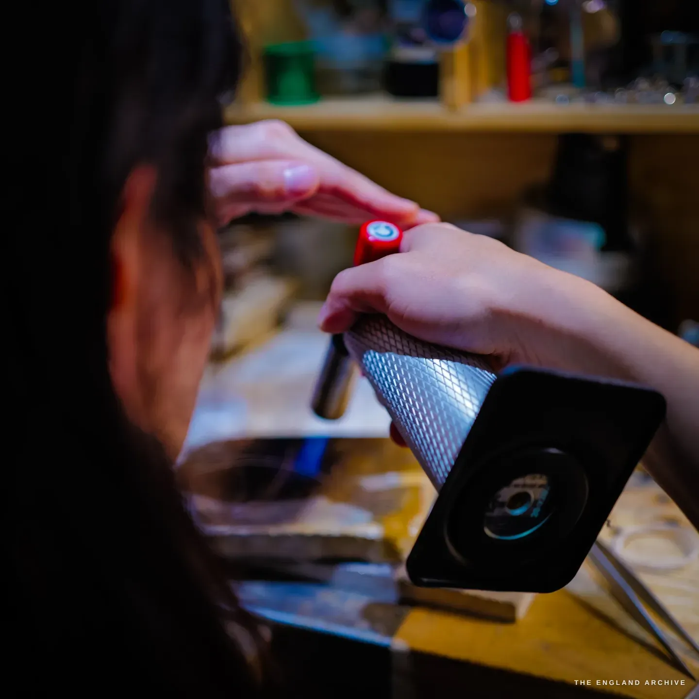 Over-the-shoulder of Yuyi filing a small workpiece against a knurled steel mandrel, a shelf of spools behind.