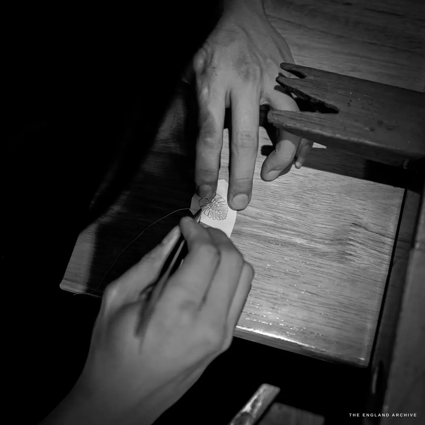 Top-down view of two hands placing a small paper monstera-leaf template on the wooden bench peg, a fine silver wire trailing from one hand.