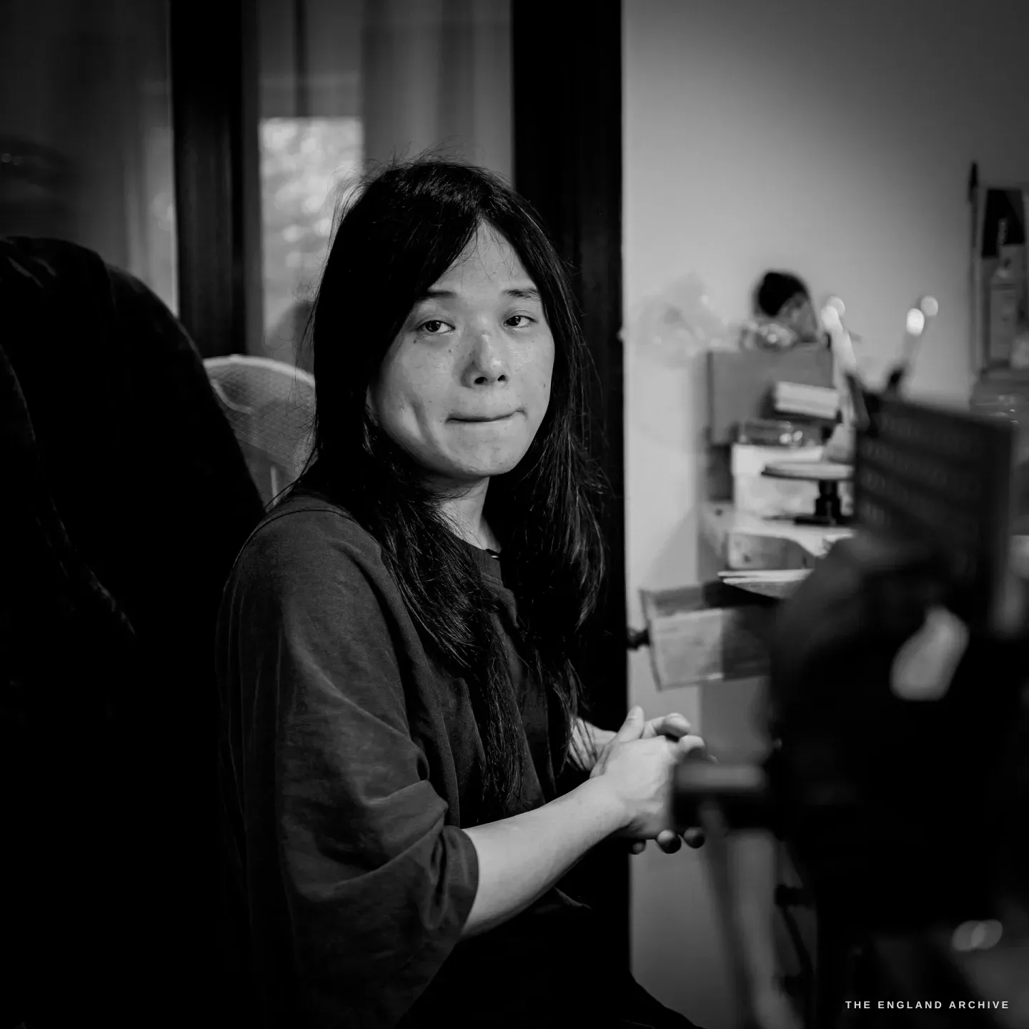 Yuyi seated at the bench, turned to the camera, hands resting on the bench pin, a window and a second figure blurred behind.